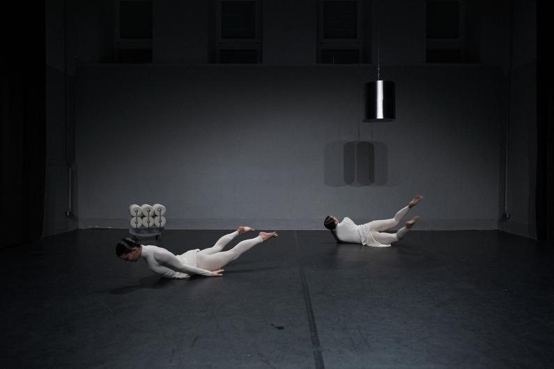 Two people in white pleated costumes move in sync on a dark stage. Their legs are bent, arms extended, with fog and spotlight creating an atmospheric tension.