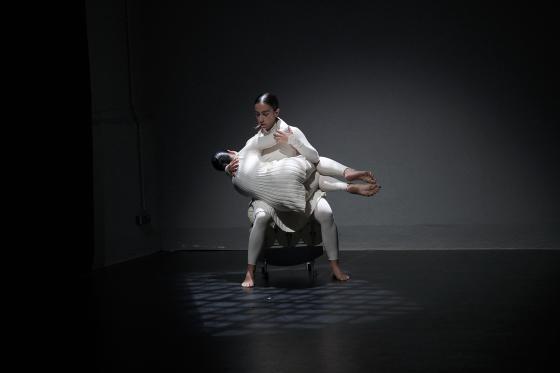 Two people lean diagonally backward in mirrored positions, their bodies taut. They wear light, form-fitting costumes as stage light cuts through the fog.