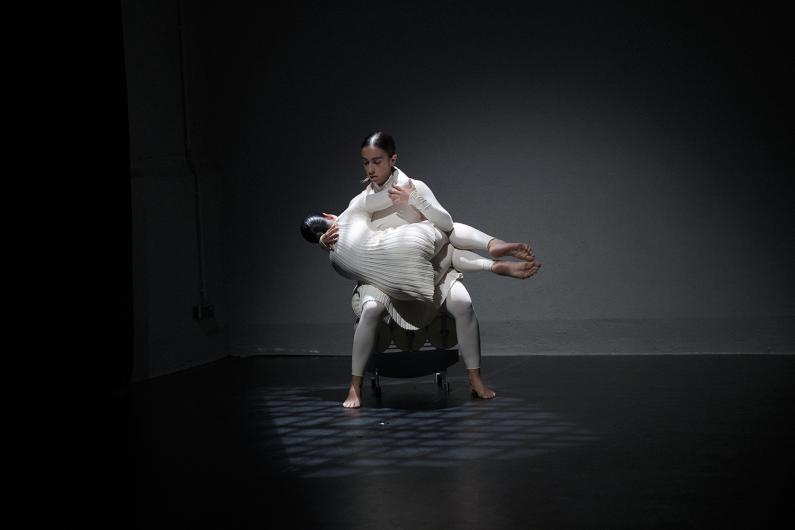 Two people lean diagonally backward in mirrored positions, their bodies taut. They wear light, form-fitting costumes as stage light cuts through the fog.