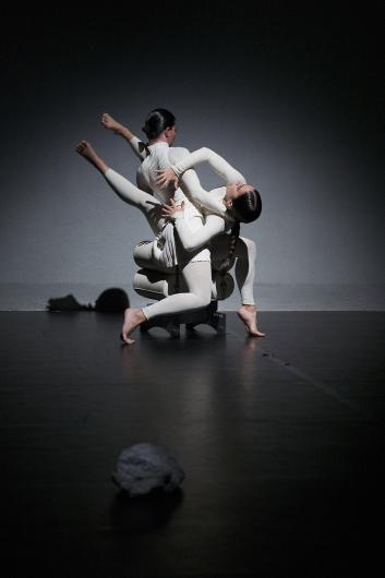 Eine Person in einem weißen, plissierten Kostüm hebt ein Bein an und streckt die Arme nach oben. Das Bühnenlicht fällt von oben, der Hintergrund bleibt im Dunkeln. 