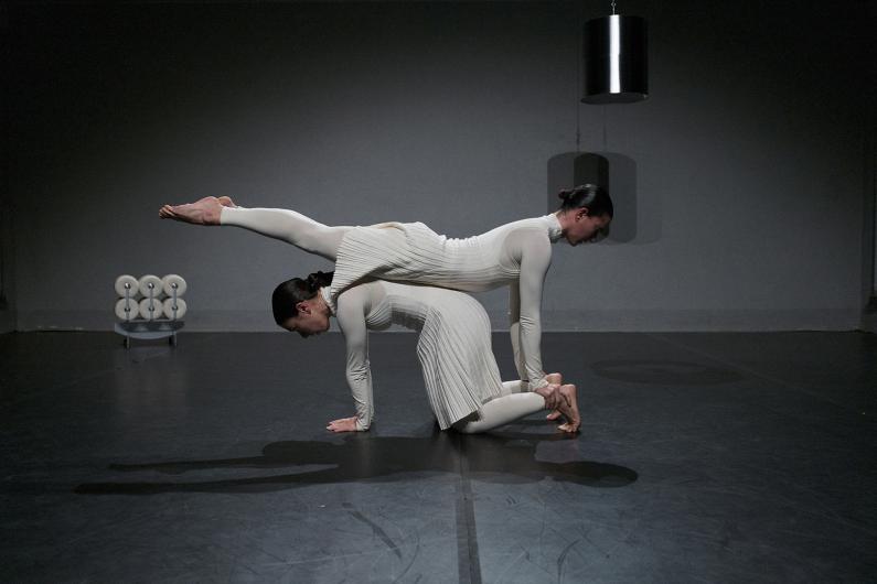 Two people lie side by side on the floor, slightly lifted as they raise their legs in the air. The scene is minimalist, with a gray wall and a hanging lamp.