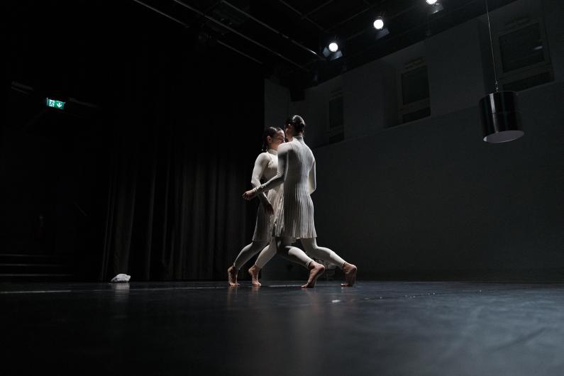 A person sits on a stool cradling another in their arms, as if asleep or carried. A spotlight highlights them against the dark stage.