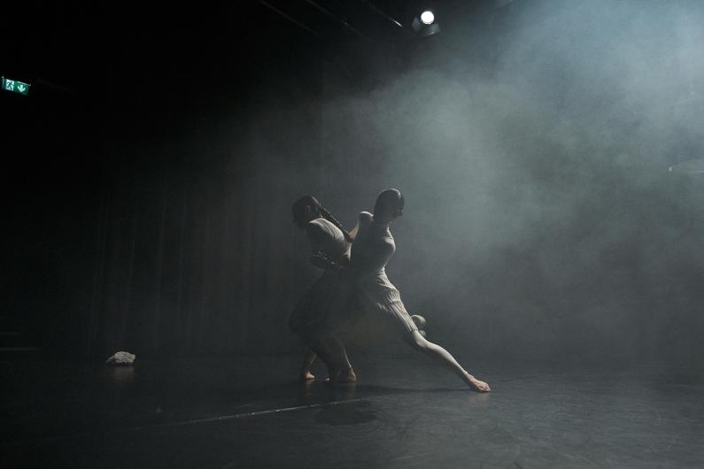 Two people in white costumes intertwine in a sculptural movement on a stool. Focused light illuminates their bodies while the rest of the stage stays dark.