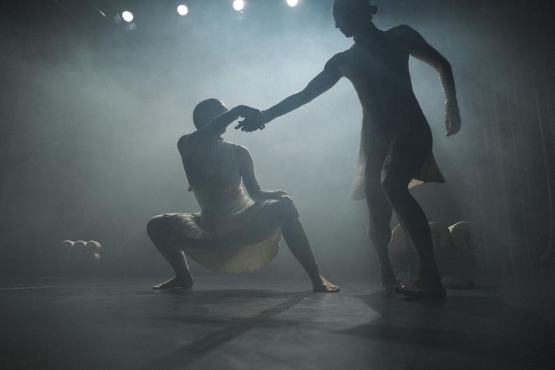 One person kneels on hands and knees while another lies horizontally across their back. Their bodies form a geometric composition.