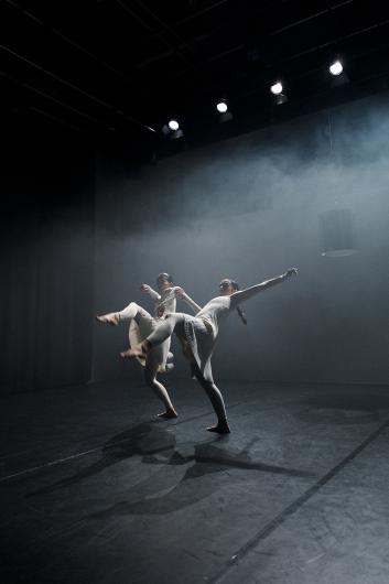 Two people stand facing each other on stage, almost touching, as light from above highlights their bodies. The scene feels intimate and calm.