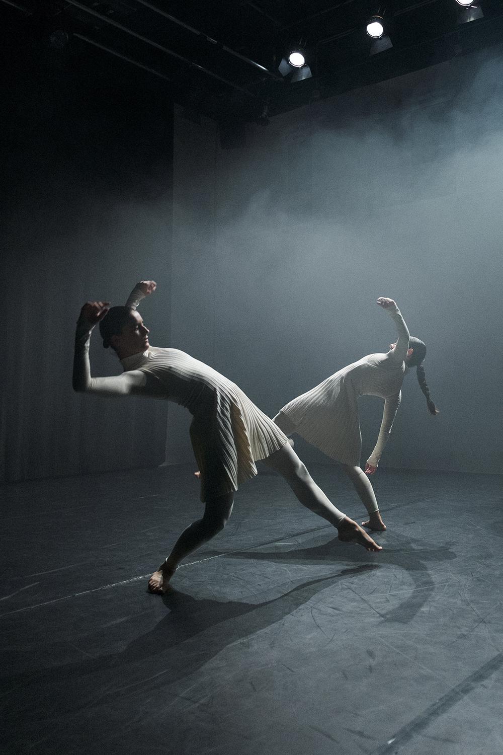 Two dancers on a dark stage lean backward in sync, arms bent and raised. They wear cream-colored pleated costumes with long sleeves and fitted tights. The space is filled with soft, misty light.