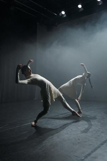 Two dancers on a dark stage lean backward in sync, arms bent and raised. They wear cream-colored pleated costumes with long sleeves and fitted tights. The space is filled with soft, misty light.