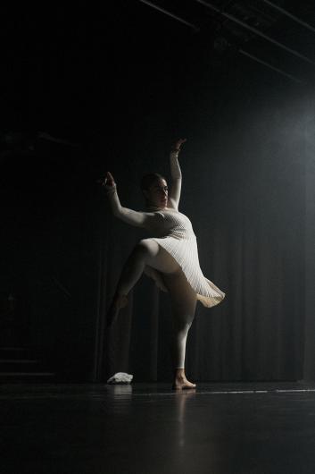 One person holds the hand of another who crouches low. Both wear light costumes, with mist and stage light enveloping their movement.