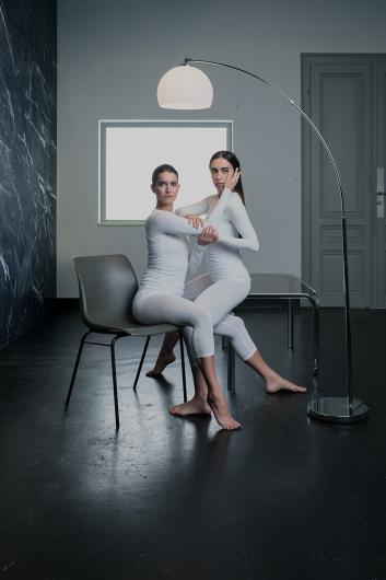 Two people in white full-body outfits sit barefoot close together. One sits on a chair, the other partly on their lap. They hold each other’s arms and face, while a floor lamp arches above them.