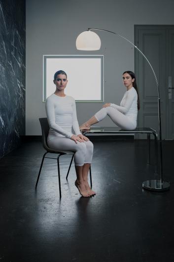 One person sits barefoot on a chair, another sits on a glass table in the background. Both wear white outfits and look seriously at the camera. A large floor lamp lights the scene.