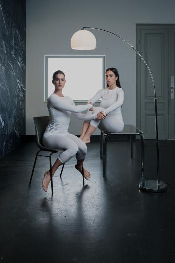 Two people in white outfits sit barefoot, one on a chair and the other on a table. They grasp each other’s forearms and maintain eye contact.