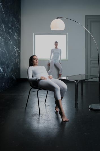 One person reclines barefoot on a chair, while the other stands framed by the window in the background. Both wear white outfits and face forward.