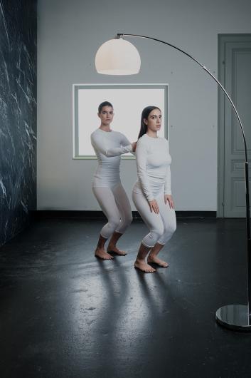 Two people in white outfits stand barefoot facing forward. One holds the other’s shoulders from behind, both look serious.