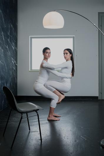 Two people embrace closely in a crouched position, barefoot and wearing white outfits. They look directly at the camera. A chair stands empty beside them.
