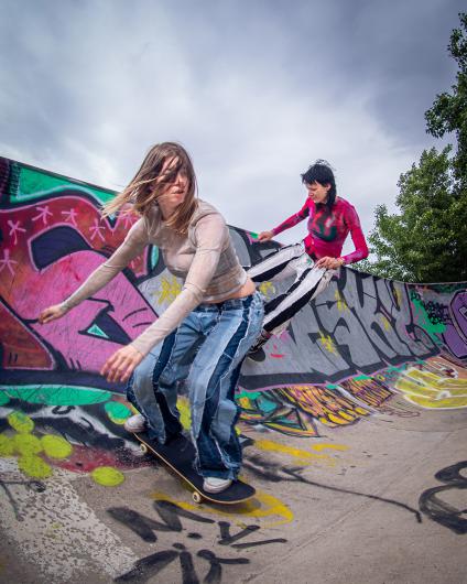 Zwei Personen in bunter Kleidung befinden sich in einer Halfpipe mit Graffiti. Eine Person fährt auf einem Skateboard, während die andere am Rand der Rampe steht und sich vorbeugt. Der Himmel ist bewölkt, Bäume sind im Hintergrund sichtbar.