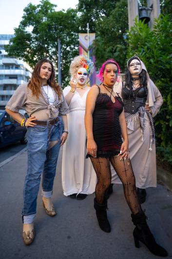 Four people in bold and diverse outfits pose confidently on a street. One person wears jeans, a tied shirt, and ankle boots. Another person wears a long white dress, a colorful wig, and has a white-painted face. The third person wears a short black dress with fishnet tights and has pink hair. The fourth person wears a corset with a long robe and face makeup styled like a nun. Trees, a car, and buildings are visible in the background.“