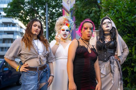 Four people in bold and diverse outfits pose confidently on a street. One person wears jeans, a tied shirt, and ankle boots. Another person wears a long white dress, a colorful wig, and has a white-painted face. The third person wears a short black dress with fishnet tights and has pink hair. The fourth person wears a corset with a long robe and face makeup styled like a nun. Trees, a car, and buildings are visible in the background.