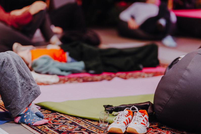 Several people are sitting or lying relaxed on colorful mats and rugs in a room. In the foreground, there are sports shoes, a small glass, and a rolled-up jacket. The atmosphere appears calm and cozy.