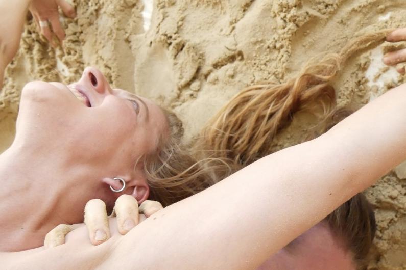 Eine Person liegt lachend im Sand, mit ausgestreckten Armen und langen Haaren, die am Boden verteilt sind. Eine Hand greift sanft an die Schulter der Person.