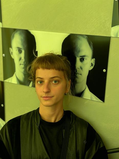 Eine Person mit kurzem, hellbraunem Pony und zurückgebundenen Haaren steht vor einer Wand mit Fotografien von Gesichtern. Die Person trägt dunkle Kleidung und blickt direkt in die Kamera.