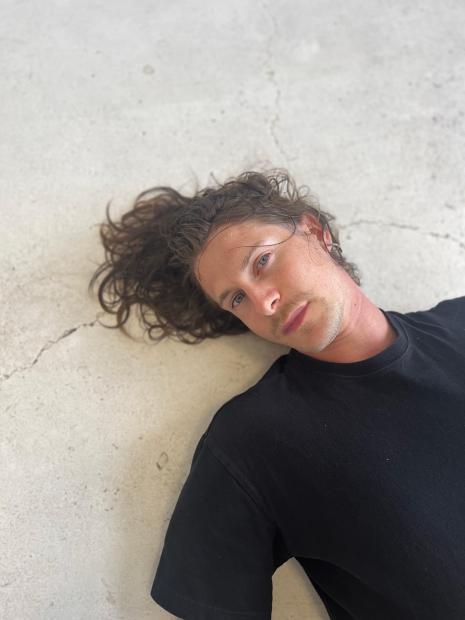 Eine Person mit langen lockigen Haaren liegt auf einem hellen Boden. Die Person trägt ein dunkles T-Shirt und schaut mit entspanntem Gesichtsausdruck in die Kamera.