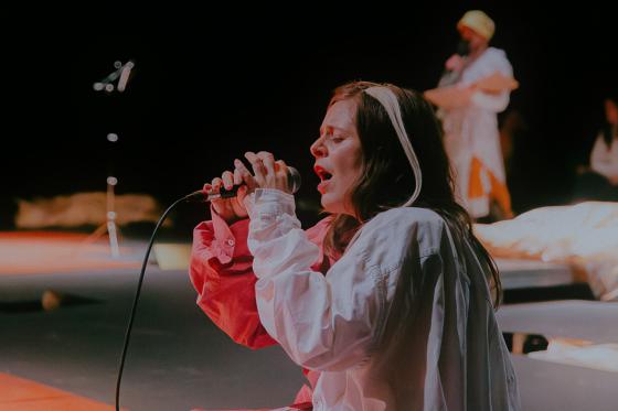 A performer sits on stage, singing into a microphone with eyes closed. They wear a two-toned blouse in white and red, appearing focused and emotional. In the background, another performer is visible, slightly out of focus, also dressed in light-colored clothing.