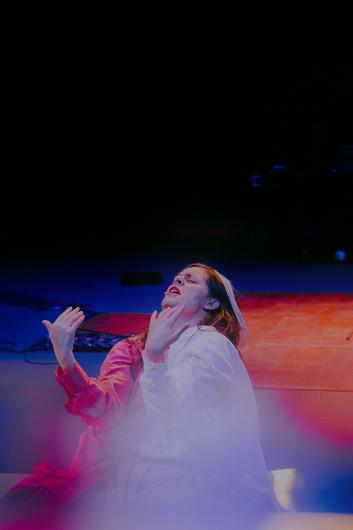 A person wearing red and white garments sits on the floor, raising both arms expressively. Their face shows strong emotion, the stage is bathed in warm light.