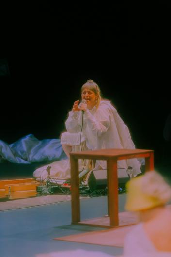 A person sits on the floor holding a microphone, singing or speaking with eyes closed. The stage is warmly lit, with technical equipment beside them.