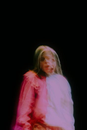 Ein verschwommenes Doppelbild zeigt eine Person in Bewegung, bekleidet mit einem roten und weißen Gewand, vor dunklem Hintergrund.