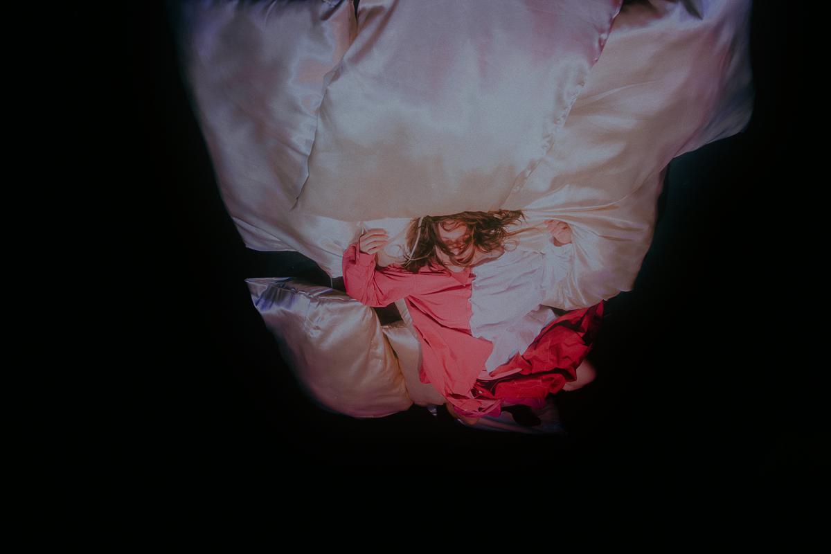 A performer kneels at the front of the stage with arms outstretched and an expressive face, as if singing or calling out. In the background, another performer lies on the floor next to a music stand. The lighting bathes the scene in strong blue and red tones, while the audience sits in semi-darkness.