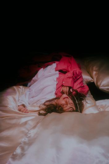 A person lies relaxed on shiny, light-colored fabric. The lighting emphasizes the soft textures, and their face appears peaceful.