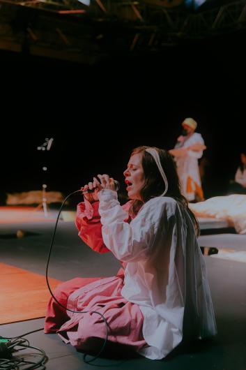A person kneels on stage holding a microphone, singing with closed eyes. Another person stands in the background under warm stage lighting.