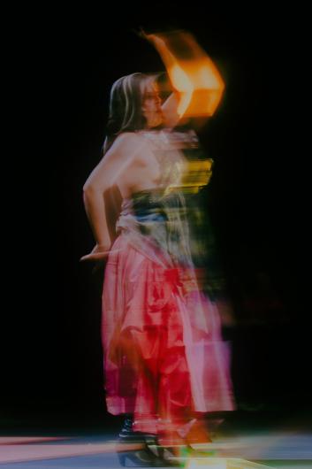 A person dances in fluid motion on a dark stage, with colorful light streaks and motion blur surrounding them.