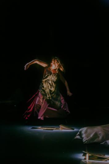 A person stands on stage in a dynamic pose with one arm extended, as light reflections fall across the floor.