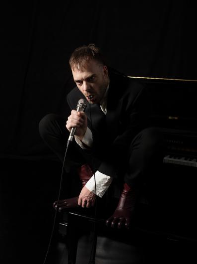 Eine Person in schwarzem Anzug und weißen Manschetten hockt auf einem Klavier, hält ein Mikrofon im Mund und blickt intensiv nach vorne.