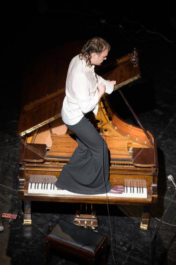 A person wearing a white shirt and wide black trousers stands barefoot on the keys of an open grand piano, singing into a microphone.