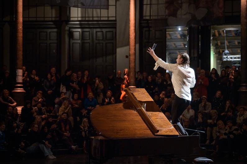 A person in a white shirt stands on a grand piano, raising a glass into the air as the audience applauds.