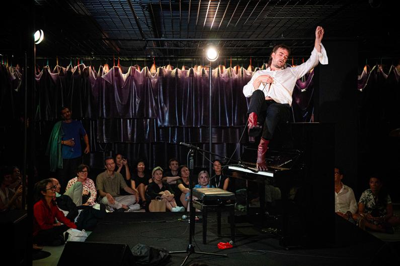 A person wearing a white shirt and red shoes sits sideways on a piano, performing expressively in front of a seated audience.