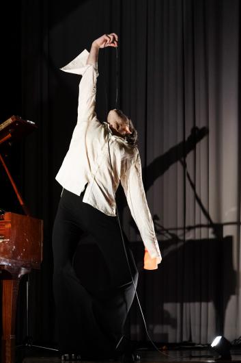 A person in a white shirt and wide black trousers leans back dramatically, holding a microphone upwards while standing next to a piano.