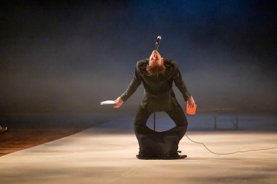 A person in a black suit bends with their head tilted back into a microphone, holding a sheet of paper in one hand and a bright orange object in the other.