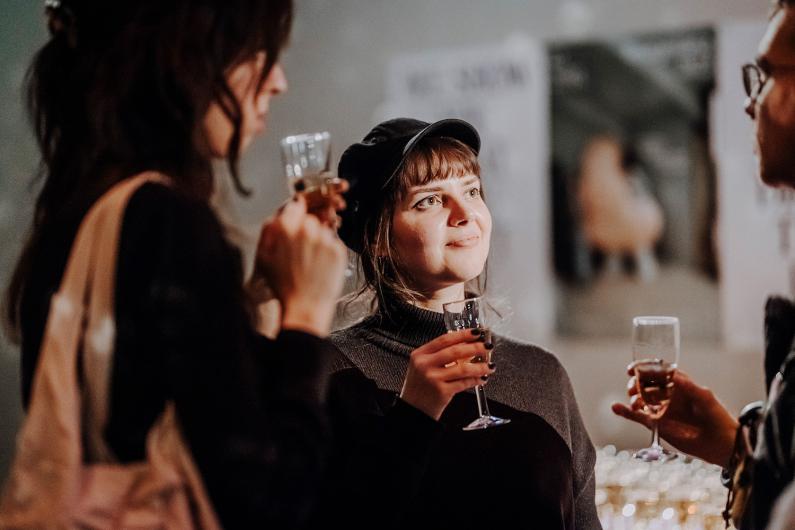 Drei Personen stehen zusammen bei einer Veranstaltung und halten Gläser mit Sekt oder Wein. Eine Person in der Mitte trägt eine dunkle Mütze und blickt lächelnd nach vorne, während mit den anderen angestoßen wird.