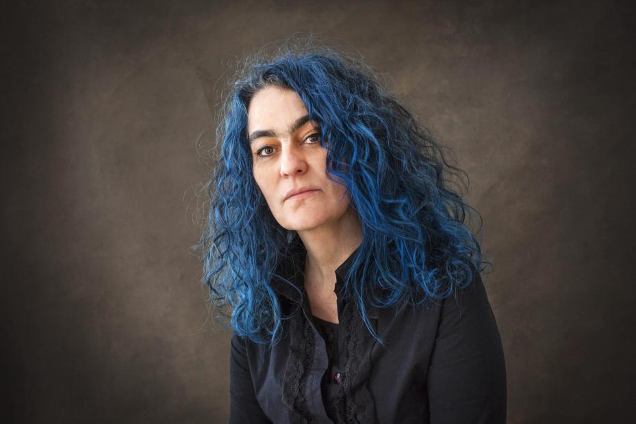 Portrait of a woman with long, curly blue hair, wearing a dark top, sitting against a neutral background and looking directly at the camera.