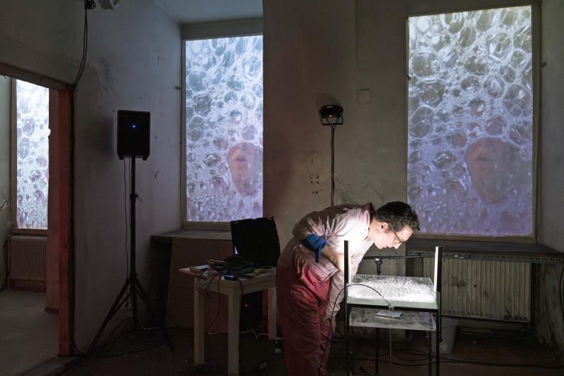 A person dressed in pink leans over a table with a lit container filled with bubbles. Behind them, large projections of faces behind soap bubbles are displayed on two windows.