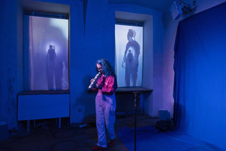 A person with long hair plays a melodica in a room illuminated with blue light. Behind them, silhouettes of other people are visible on two large window screens.