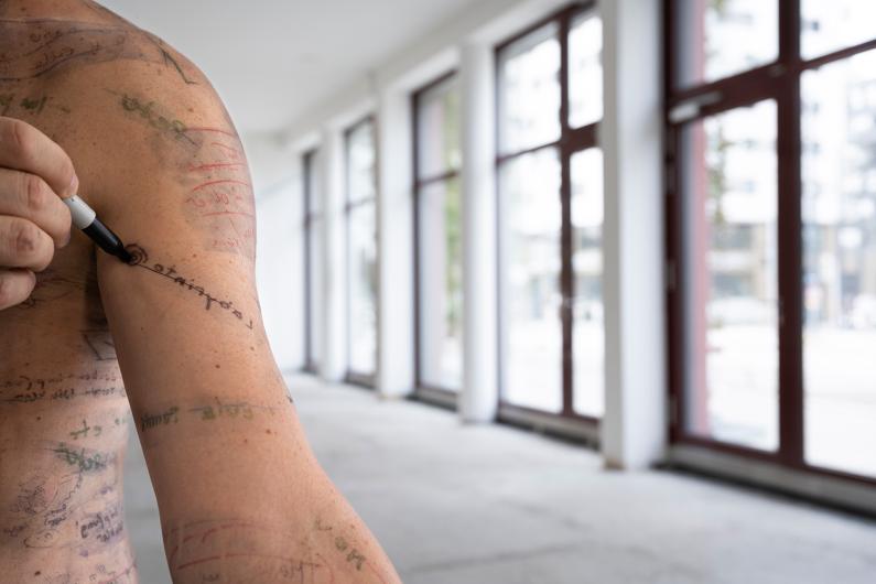 A bare upper arm covered with handwritten notes and drawings. A hand holds a black marker, writing on the skin. In the background, a bright empty room with large windows.