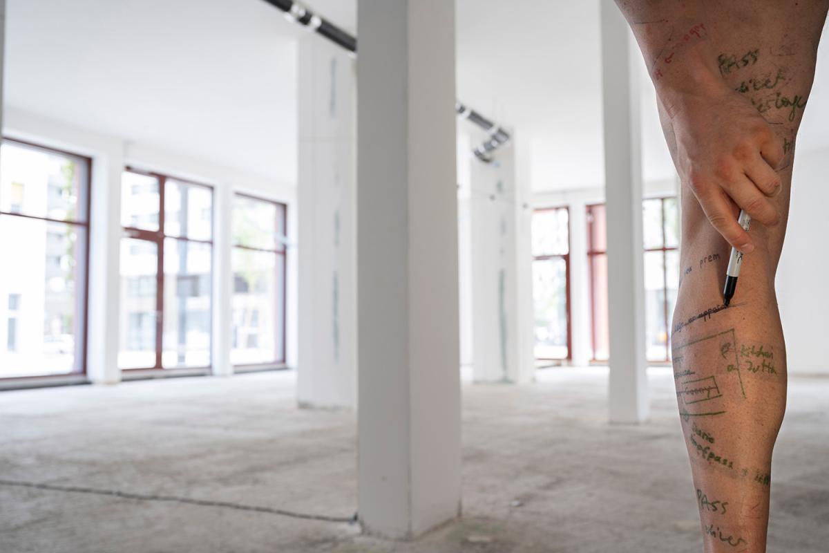 A bare leg with handwritten notes and drawings on the skin. A hand applies a black marker. In the background, an empty bright room with columns and large windows.