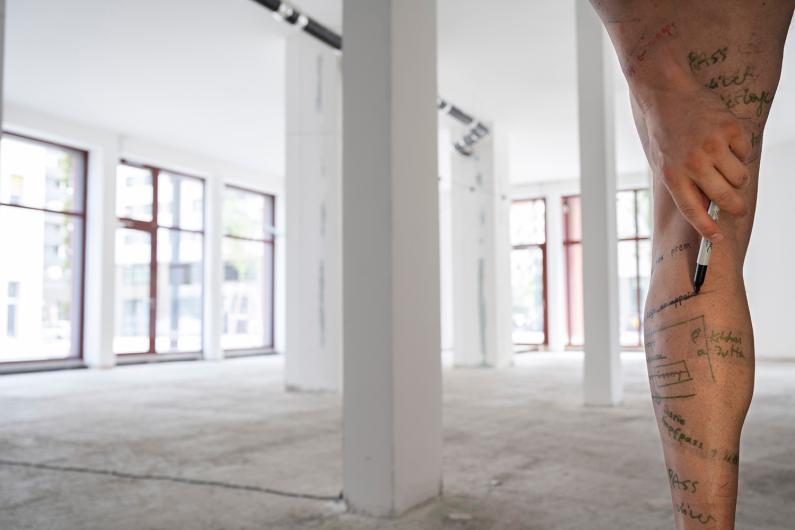 A bare leg with handwritten notes and drawings on the skin. A hand applies a black marker. In the background, an empty bright room with columns and large windows.