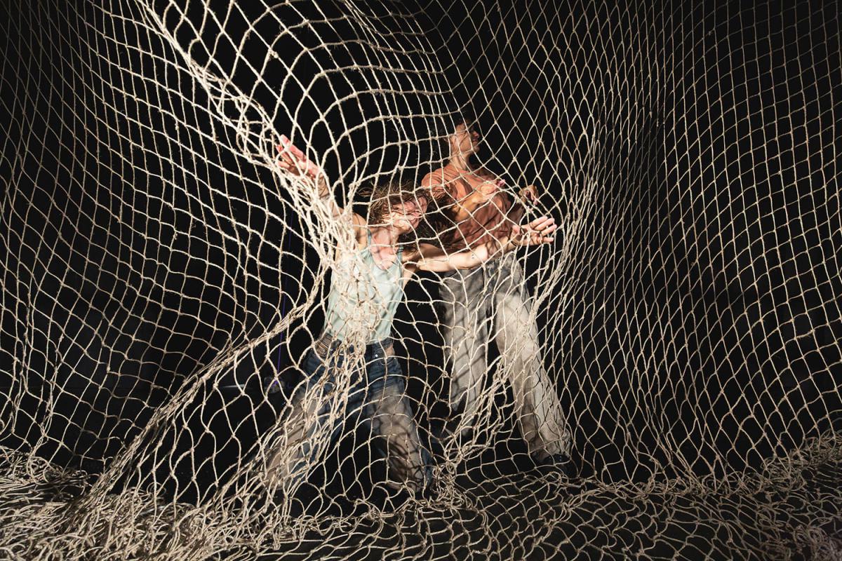 Two people move behind and within a large net made of thick ropes. Their bodies are partly obscured by the net as they stretch their arms and upper bodies outward in dynamic poses. The scene is dramatically lit against a black background.