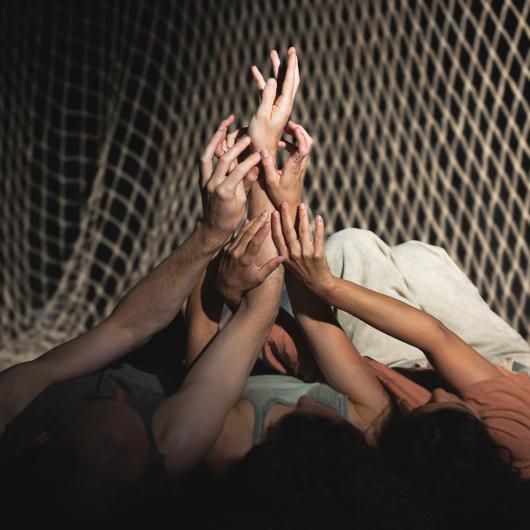 Several people lie close together on the floor under a net, with their hands reaching upwards together.