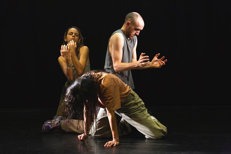 Three people dance on a dark stage. One kneels bent forward on the floor, while two stand behind with tense hands and closed eyes.